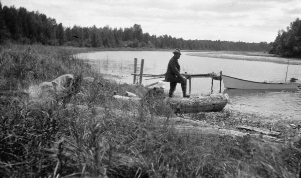 An unidentified man poses near a makeshift pier on the Mink River.  A small boat with outboard motor is tied to the pier. The photographer's son Fedy (Ferdinand Leonard), identified by his white hat, sits near a large log.