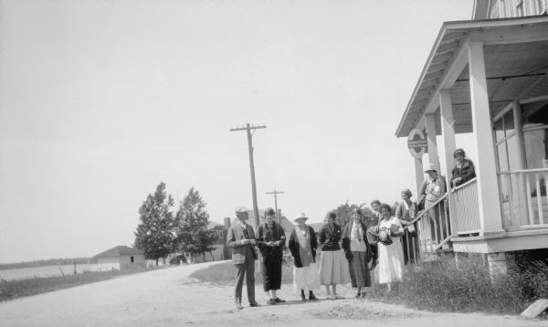 A man stands with a group of nine women posed in front of a small commercial building. A sign in the shape of a tire advertises "Goodyear Service Station." A narrow road runs along the shore of the harbor.
