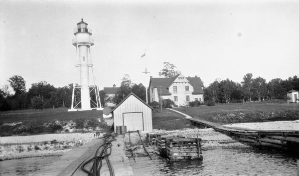 View of the Plum Island rear range lighthouse from the end of the pier. The brick house was home to the lighthouse keeper and the first and second assistant keepers. A boathouse and barn are also seen. There was also a front range lighthouse on Plum Island.