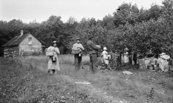 Young men and woman pose with baskets and pails, and a girl stands by her bicycle at the Hotz cherry orchard. There are baskets, lids, and boxes of cherries on the ground. An old log cabin is in the background.