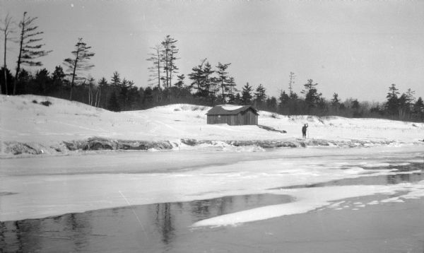 View from water of a man with a long walking stick posing near a wooden boathouse on Europe Bay. There is ice on the water near the shore.