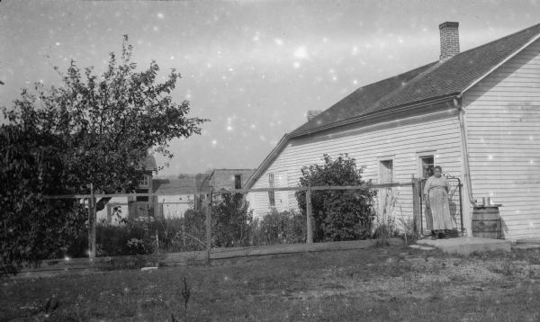 A woman stands at the gate beside a wood frame house, possibly at Newport. The downspout on the house drains into a rain barrel. There are outbuildings behind the house.