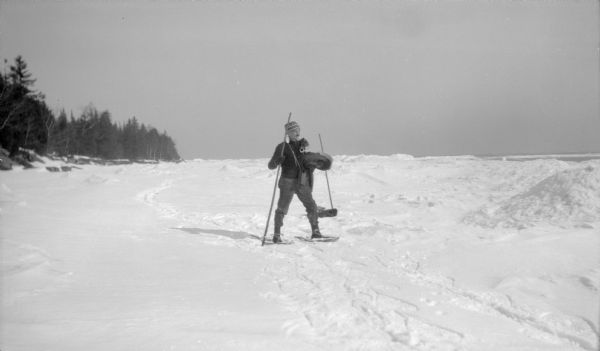 A man, likely Ferdinand Hotz, on short snowshoes carrying a long walking stick and his coat. A camera case and pair of ice skates hang from straps around his neck. Another stick is standing in the snow behind him.