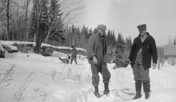 Two men hold the heads of a pair of whitetail bucks with locked antlers. There is a boy and a dog in the background near a rock outcropping and snow is on the ground. A building is in the background on the right.