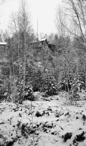 The Hotz cottage is seen from the base of the bluff. Snow covers the birch and conifer trees.
