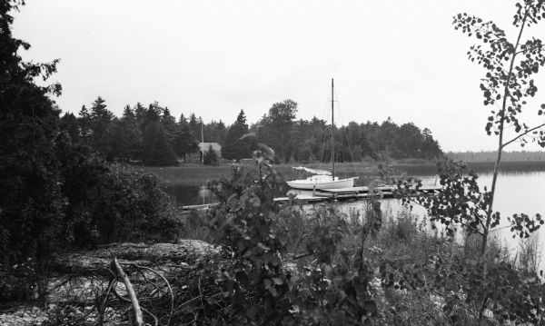 A sailboat is moored to a wooden pier at North Bay. There is an old wooden building in the background across a marsh area.