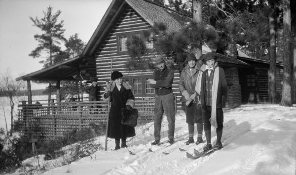 Winter scene with Ferdinand Hotz, holding a cigarette, posing on skis. His wife, Clothilde, stands on the left with a long walking stick; daughter Helen (Leni) is on the far right next to an unidentified woman. Both young women are wearing skis. The Hotz Lake Europe cottage with its massive chimney and rustic porch railing is the background. Two men stand on the porch.