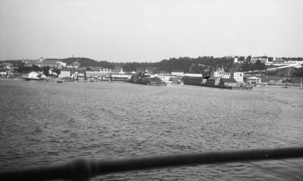 View of Mackinac Island from a boat offshore. There are two large wooden piers with warehouses, and a beach on the right. Commercial buildings line the waterfront. The Grand Hotel, with its numerous columns, stands on a hill on the left.