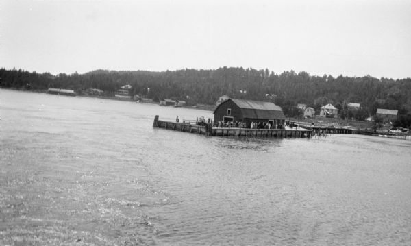 A large group stands on the pier at Fish Creek as a boat leaves. A barn-like warehouse stands on the pier; houses, boathouses, and outbuildings are visible onshore.