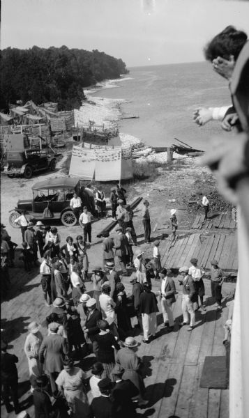 Passengers lean on the railing of a boat at a dock as a well-dressed crowd waits below. There are two cars parked nearby. Fishing nets dry on net rollers along the shore. A rocky beach stretches toward the horizon.