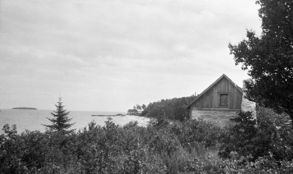 View of rustic cabin with a large stone chimney standing by the shore. There is another house or boathouse in the far background near the remains of a pier. One of the Strawberry Islands is seen on the horizon.