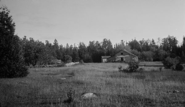 Two deteriorating log buildings stand in a stony meadow. There is a stone fence on the left and trees beyond.