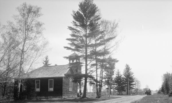 Three women pose on the steps of the Baptist Church at Ellison Bay, which has an open bell tower. A car is parked a short distance down the dirt road.