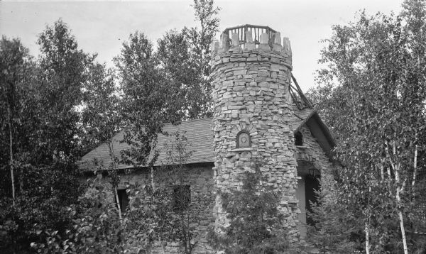 The stone garage and attached tower at the Hotz cottages at the top of Fish Creek Hill. The tower features a stone parapet with wooden railing, as well as a small, arched, stained glass window. There appears to be a ladder leading up to the tower from the roof of the garage.