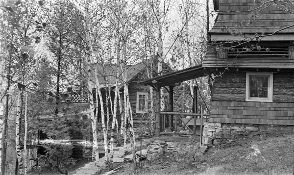 The pumphouse, foreground, and cottage at the Hotz compound at the top of Fish Creek Hill. Also shown is the log outhouse, left. The buildings are clad in logs and shingles, with rustic railings and details. There was a tank above the pumphouse, which has since been rebuilt closer to Highway 42.