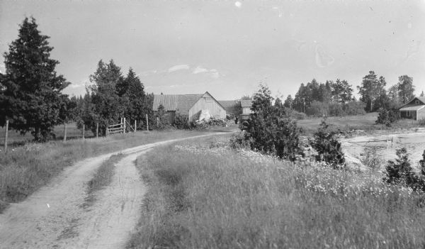 View down dirt road leading to the Carlson farm at Nelson Bay in Peninsula State Park. A man stands in the background among the farm buildings near a parked car. The Lazaar cottage is at far right.