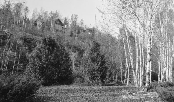 View of the Hotz family compound of cottages from below the bluff at Fish Creek. A flag waves in the breeze; there are birch trees and conifers in the foreground.