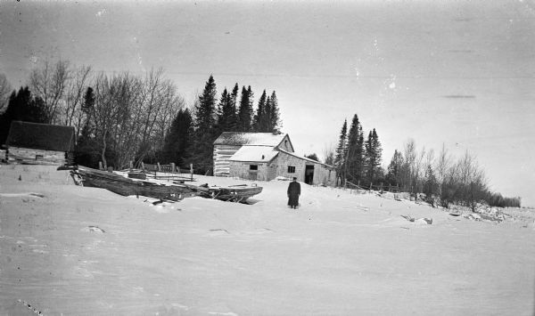 An unidentified man poses near a wrecked boat. A log house with two lean-to additions is behind him and there is a smaller log building on the left. Snow covers the ground and shoreline.