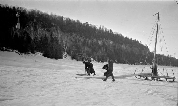 Three men work with an iceboat just offshore near Cottage Row Road.  The sail and boom lie on the ice.  There are boathouses and three windmills along the shore; a cottage is visible through the trees. A bluff rises just behind the snow drifted shore.