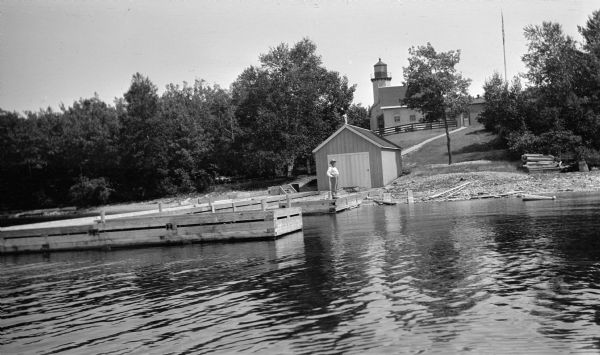 View from water of a man with a beard standing on a pier near a small boathouse. In the background is a lighthouse with keeper's house and outbuilding.