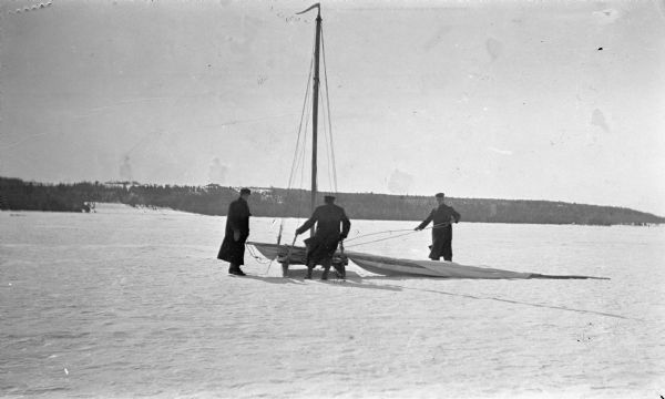 Three men work with an iceboat on Lake Michigan. The sail and boom lie on the ice.