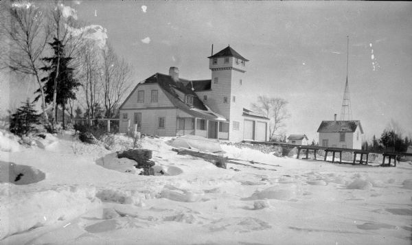 Winter view of the Coast Guard Life Saving Station at Baileys Harbor. Snow has drifted on the shore, where a small boat rests.