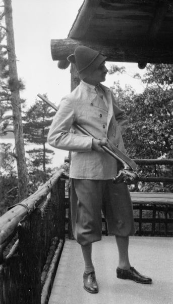 Ferdinand Hotz poses in Bavarian dress, holding a carved walking stick, on the porch of his cottage on Europe Lake. He wears a felt hat with braid and a large brush. The porch has a rustic railing.
