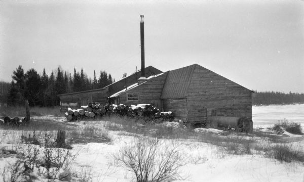 Logs are stacked beside a sawmill in a winter landscape. The mill has a tall smokestack and stands near the shore.