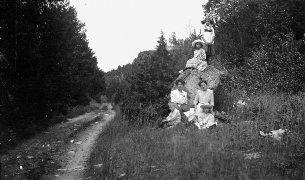The photographer's wife, Clothilde, sits, (right) in front of a rock with an unidentified woman, as daughters Alice, standing, and Helen pose on top of the rock; son Ferdinand Leonard, left, and daughter Margaret sit in front of them on the grass. A one-lane dirt road leads further into the woods.