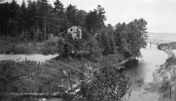 Elevated view of a two-story house sited beside Shivering Sands Creek where it enters Lake Michigan. There is a rustic footbridge over the stream close to the shoreline.