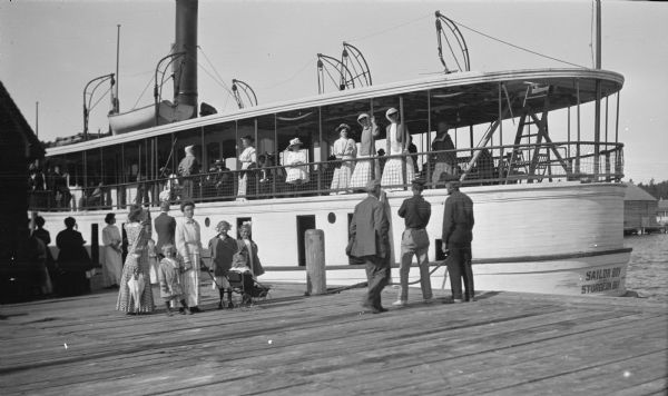 The photographer's family poses on the dock beside the ship "Sailor Boy" of Sturgeon Bay. Clothilde Hotz holds the hand of Ferdinand Leonard (Fedy); daughter Margaret is in the stroller; daughters Alice and Helen are on the right. Other well-dressed men and women stand on the dock and on board the ship.