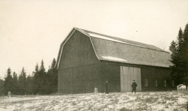 Two men pose in front of a large barn at Newport Village.  The barn was used to store lumber.