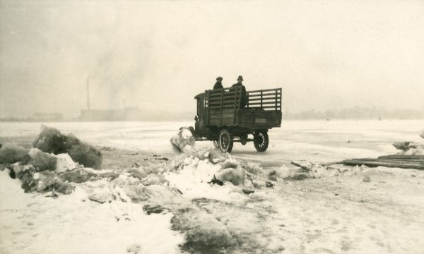 Two men stand in the open back of a pickup truck.  The truck, equipped with chains on the rear tires, has driven down a wooden ramp onto the ice.  There is a mill or factory with tall smokestack on the far shore and ice fishing shanties on the right.