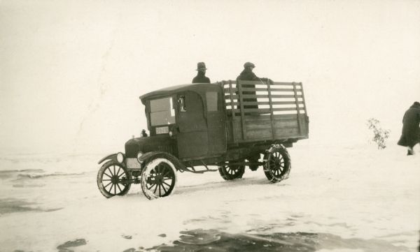 Two men stand in the open bed of a pickup truck as a third person walks away. The truck, equipped with snow tires, is parked on the ice just offshore.