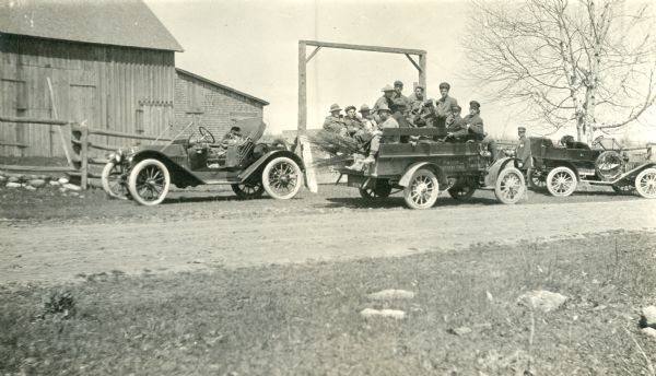 A pickup truck carrying a crew of 12 men is parked between two automobiles in front of a barn. The truck is identified as "Bingham & Lawrence Fruit and Nursery Stock." There are bundles of young trees in the truck. One man at the rear smokes a pipe; several men smoke cigars.