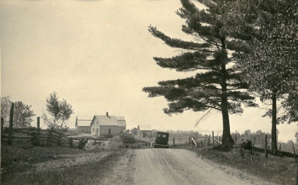 A car is parked along a narrow dirt road with a house, barn and outbuildings in the background. A split-rail fence lines the left side of the road. There is a far view of a bay.