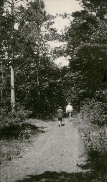 Two men walk away from the camera, down a narrow country road. Both are wearing knickers and knee socks; the man on the left wears a hat and carries a walking stick.