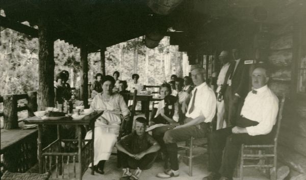 A group poses on the rustic porch of the Hotz family cottage on Europe Lake. There are dishes and food on the tables. The photographer's wife, Clothilde, with glasses, faces the camera on the left; son Ferdinand Leonard (Fedy) sits on the porch floor and daughter Alice holds a dachshund.