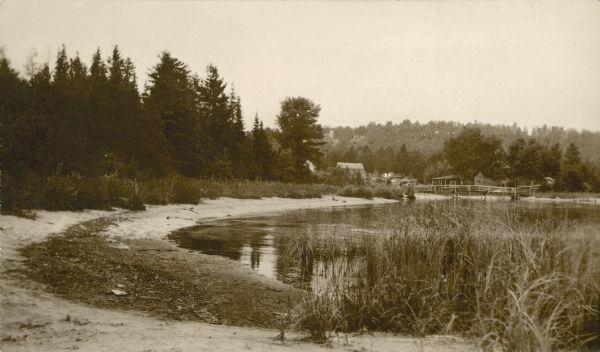 View along shoreline towards a rustic foot bridge which crosses the mouth of Fish Creek, which separates the village of Fish Creek from Peninsula State Park. In the far distance, the Hotz family compound with stone tower can be see on the bluff.