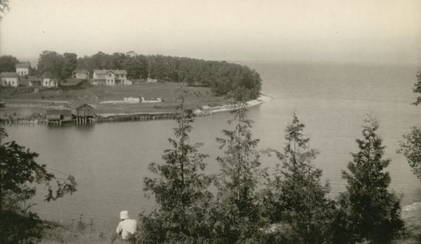 Elevated view of the photographer's son, Ferdinand Leonard (Fedy) sitting on the bluff overlooking Fish Creek Harbor. The old warehouses are seen on the pier; empty stone foundations stand near the shore with other houses beyond.