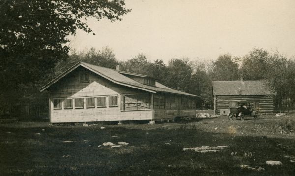 The Hotz Gibraltar Orchard cherry camp house with a low dormer in front; all windows and the porch are shuttered. There is a car parked in front of a log cabin in the background.  Ski View Drive, near its intersection with Highway 42, now occupies the site of the cherry camp house.