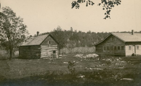 Looking southeast from behind the camp house, right, and log cabin, left, at the Gibraltar Orchard cherry camp. All the windows on the cottage are boarded up. A hill rises in the background beyond the blooming cherry trees.  Ski View Drive, near its intersection with Highway 42, now occupies the site of the cherry camp house.