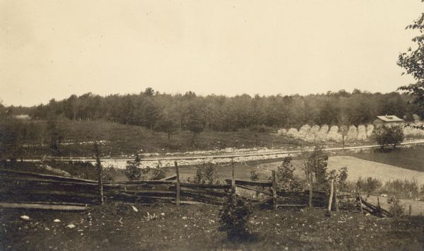 Elevated view across Sturgeon Bay Road, now Highway 42, toward the Gibraltar Orchard cherry camp. There is a wooden fence in the foreground, and the camp cottage is visible on the right.