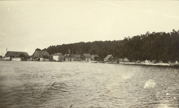 View from the water of Washington Harbor on Washington Island. There are boats moored and warehouses along the pier. Fishing nets dry on racks on the shore.