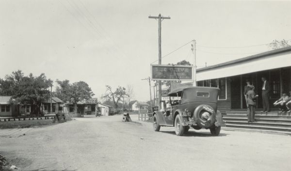 A car is parked in front of "The Olive Dining Room." Several men sit or stand on the porch steps. There is a Coca-Cola sign on a small building in the background, and another sign advertises "Drugs".