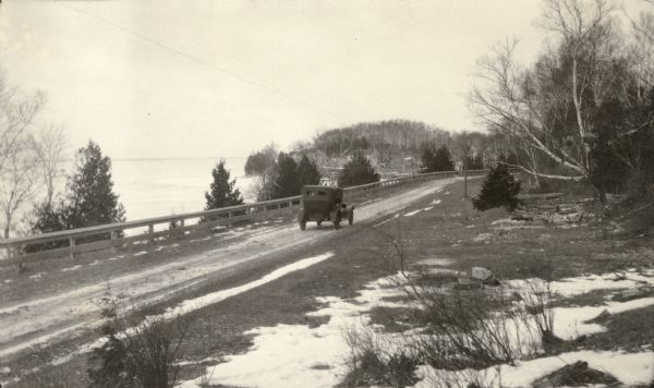 A car travels down a narrow road along the shoreline towards the abandoned town of Fayette. There are patches of snow on the ground. Houses are visible in the background.