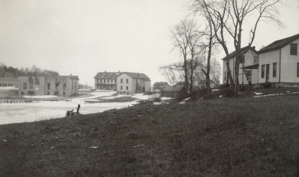 There is smoke damage to the bricks evident above the windows of the company store in this view of the ghost town of Fayette. To the right, with dormers, is the hotel; next is the town hall, all on the shore of Snail Shell Harbor. A boat appears to be grounded on the shoreline below the hill in the foreground.