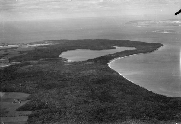 Aerial view of the northern tip of the Door Peninsula, looking north. Europe Lake is in the center, with Europe Bay to the right. Plum Island and Washington Island are visible in the right background. Cultivated fields are clearly defined on the left. Much of the wooded area south of Europe Lake was owned by Ferdinand Hotz and is now Newport State Park.
