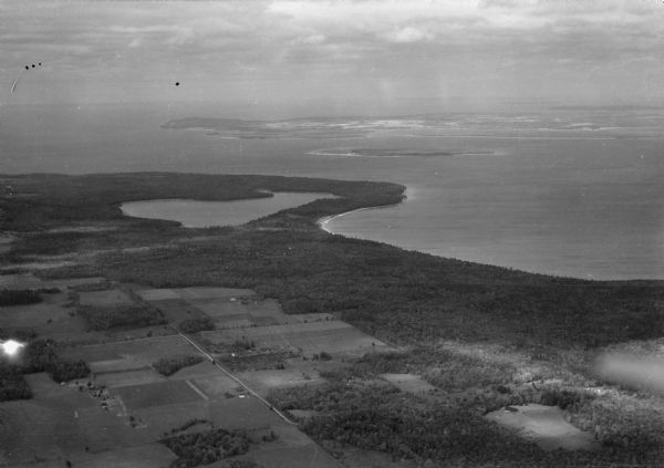 Aerial view over northern Door County, looking northeast across Europe Lake and Europe Bay toward Plum and Washington Islands. Farm fields and orchards contrast with extensive wooded areas. Most of the wooded areas south (to the right) of Europe Lake are now part of Newport State Park and at the time of the photograph were owned by Ferdinand Hotz.