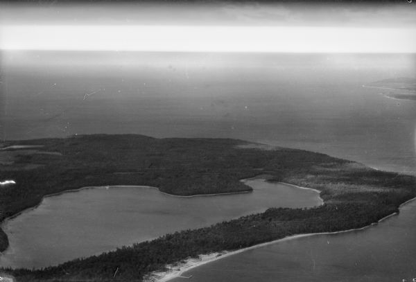 Aerial view, looking north, across the beach and pier on Europe Bay to Europe Lake and beyond to Washington Island. The land south (to the right) of Europe Lake was owned by Ferdinand Hotz at the time of this photograph and is now part of Newport State Park. The Hotz Europe Bay pier is seen extending from shore near the bottom of the image.